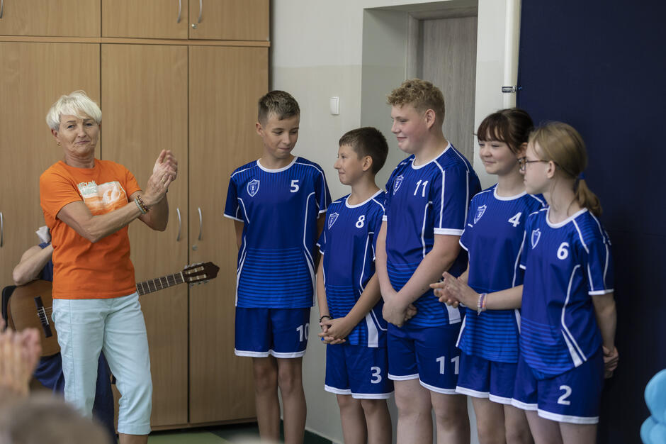Na zdjęciu grupa uczniów w niebieskich strojach sportowych stoi obok siebie, uśmiechając się i spoglądając na siebie nawzajem. Obok nich starsza kobieta w pomarańczowej koszulce i jasnych spodniach bije brawo, a w tle widać gitarzystę. 