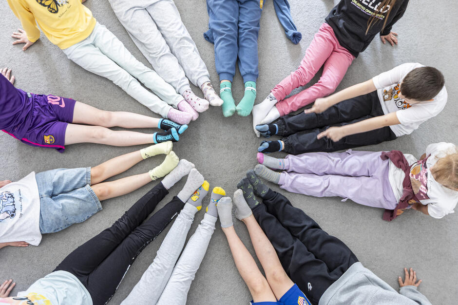 Na zdjęciu widać grupę dzieci siedzących w kręgu na podłodze, z nogami skierowanymi do środka. Mają na sobie kolorowe skarpetki, które tworzą barwny wzór wokół środka koła.
