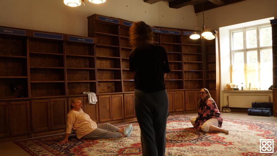 Trzy kobiety w pomieszczeniu w starym wnętrzu dawnej księgarni. Dwie kobiety siedzą na dywanie, jedna stoi. Przy ścianach stoją puste regały na książki.
