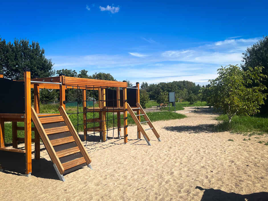 Na zdjęciu widać plac zabaw z drewnianą konstrukcją do wspinania, ustawioną na piaszczystym podłożu. W tle rozciąga się zielony teren z drzewami i ścieżkami, a nad całością rozpościera się błękitne niebo. 