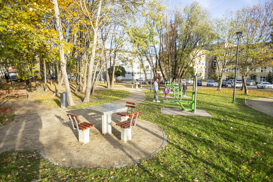 Na zdjęciu widać park z alejkami, stołem do gry z ławkami oraz zieloną siłownią plenerową, z której korzystają mieszkańcy. Otoczenie zdobią drzewa z jesiennymi liśćmi, a w tle widać budynki i zaparkowane samochody.
