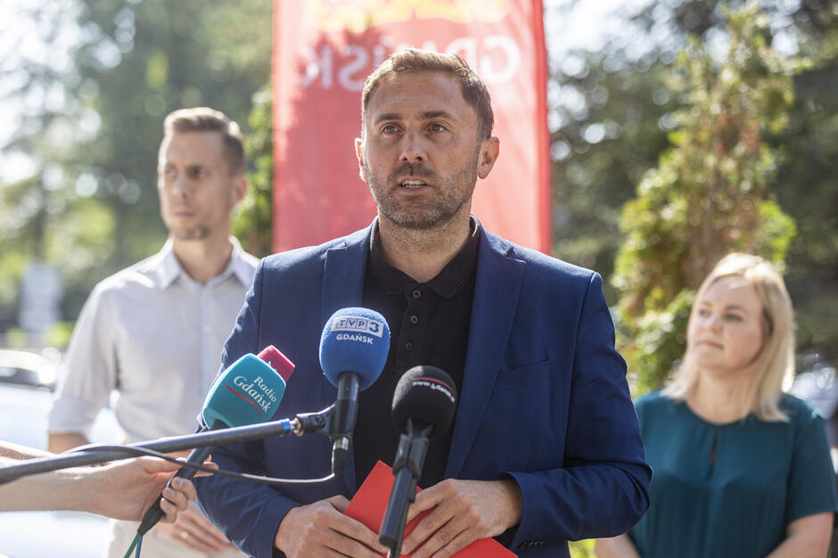 na zdjęciu mężczyzna w średnim wieku, w niebieskiej marynarce, mówi do mikrofonów