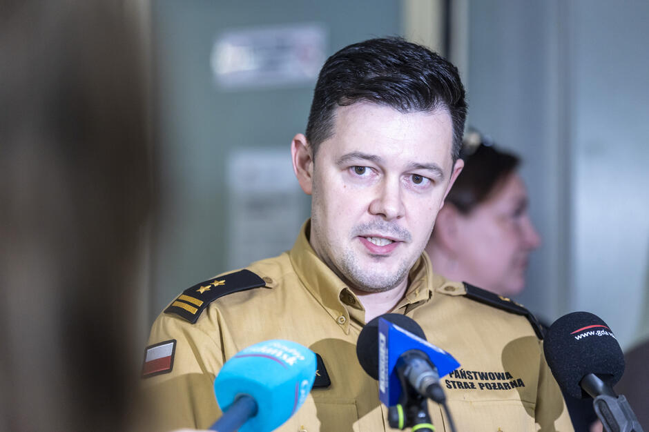 Na zdjęciu znajduje się strażak w mundurze Państwowej Straży Pożarnej, udzielający wypowiedzi mediom podczas konferencji prasowej. Przed nim ustawiono kilka mikrofonów z logotypami lokalnych stacji radiowych i telewizyjnych. 