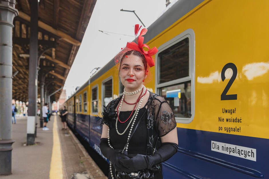 na zdjęciu kobieta stojąca przed żółto-niebieskim pociągiem na stacji kolejowej. Ma na sobie czarną sukienkę z czerwonym toczkiem na głowie. Pociąg ma numer 2 i słowa  Uwagal Nie wsiadać po sygnale 