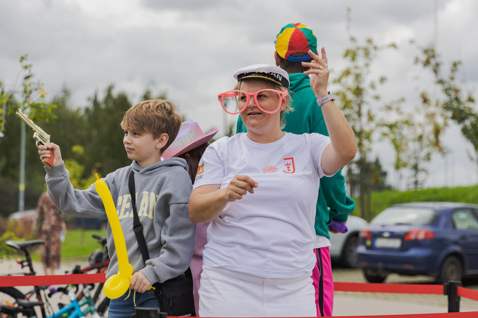 na zdjęciu kobieta w za dużych kolorowych oprawkach okularowych, obok niej stoi dwoje dzieci, bawią się