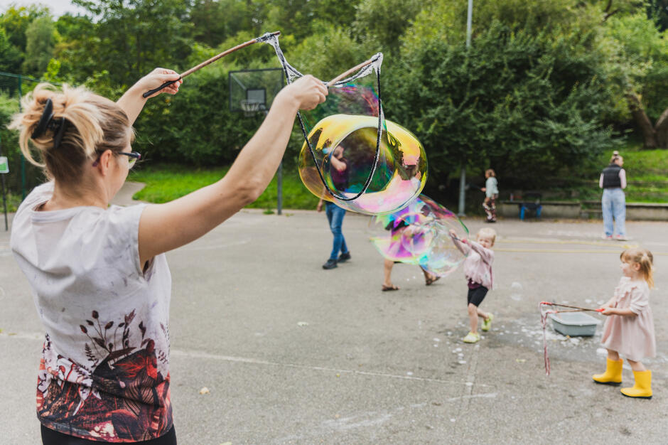na zdjęciu tyłem stojąca kobieta, trzyma dwie specjalne linki z których wypuszcza ogromną bańkę mydlaną, w tle bawiące się dzieci