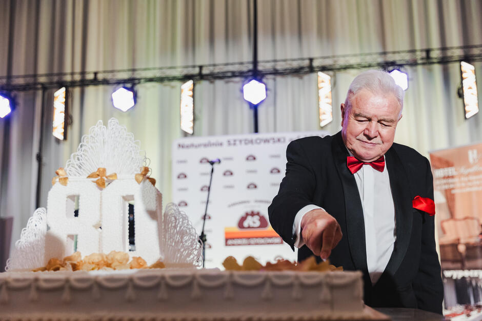 Starszy, ubrany elegancko, mężczyzna stoi po prawej stronie, kroi wielki płaski tort