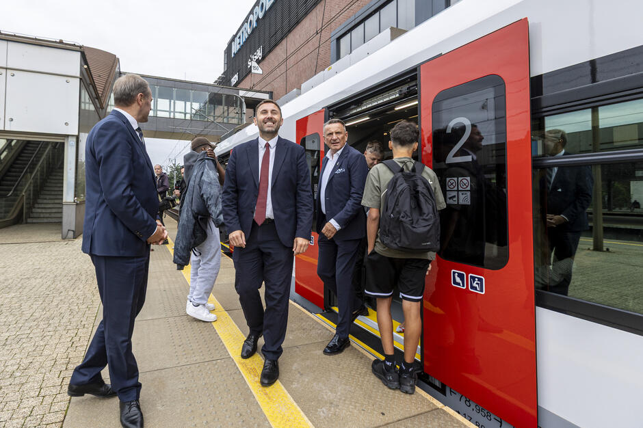 Na zdjęciu widać ludzi wysiadających z nowoczesnego pociągu na peronie – wśród nich znajdują się elegancko ubrani mężczyźni w garniturach oraz pasażerowie w codziennych strojach. W tle widoczny jest budynek z napisem „METROPOLIA” oraz kładka łącząca części dworca. 