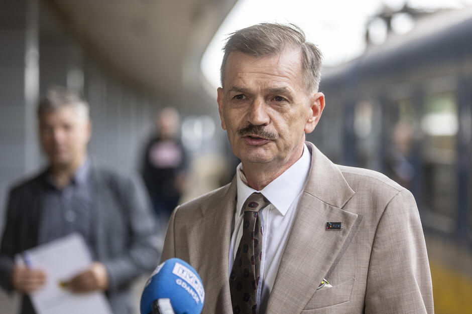 Na peronie kolejowym mężczyzna w beżowym garniturze udziela wypowiedzi do mikrofonu oznaczonego logo TVP Gdańsk. W tle widoczni są inni ludzie oraz stojący przy peronie pociąg. 