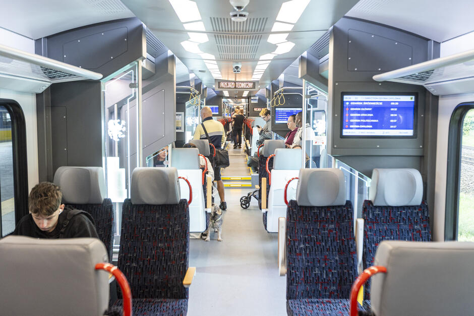Wnętrze nowoczesnego pociągu SKM jest jasne i przestronne, z wygodnymi siedzeniami i elektronicznymi tablicami informacyjnymi. W środku podróżni siedzą, korzystają z telefonów, a na korytarzu widać także psa. 