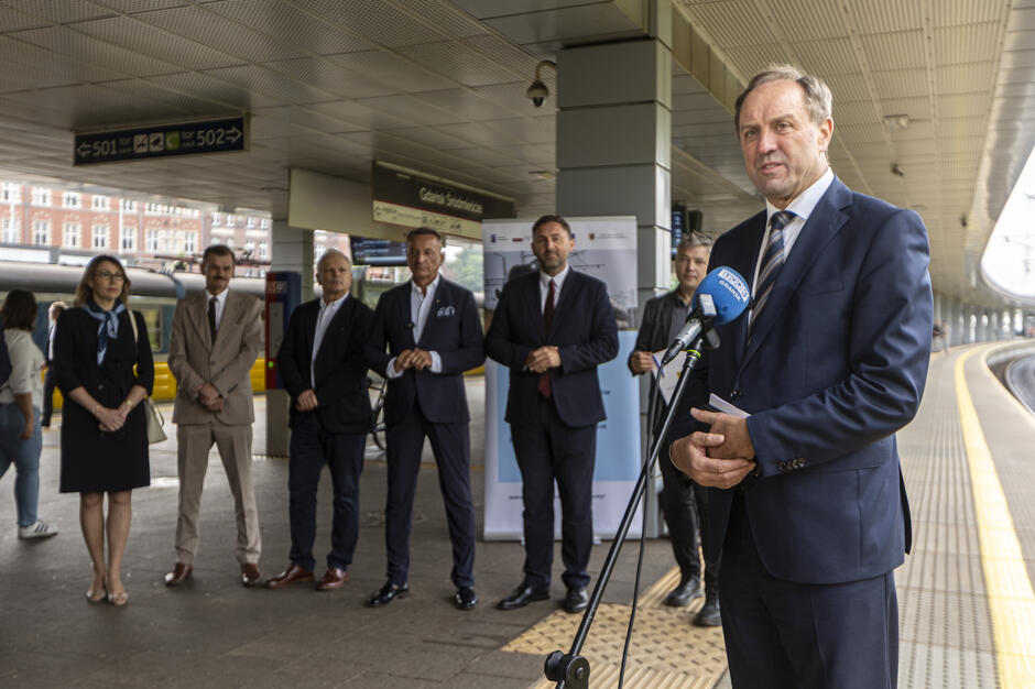 Na peronie stacji Gdańsk Śródmieście odbywa się konferencja prasowa, podczas której mężczyzna w garniturze przemawia do mikrofonu TVP Gdańsk. Za nim stoi grupa osób, również w formalnych strojach, tworzących oficjalne tło wydarzenia.
