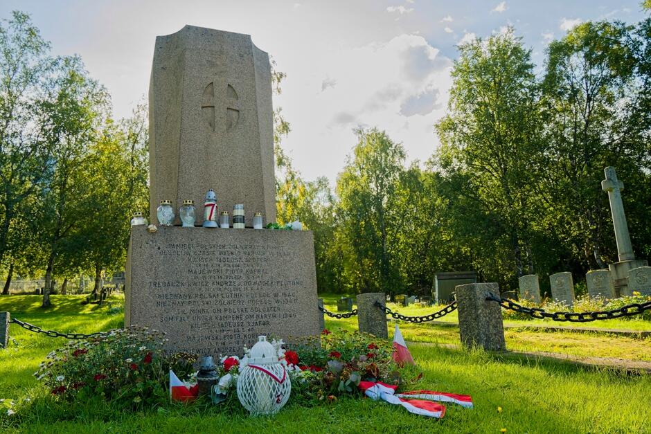 Na zdjęciu widać pomnik upamiętniający polskich żołnierzy poległych w Norwegii w 1940 roku, otoczony zniczami, kwiatami i biało-czerwonymi flagami. Monument znajduje się na cmentarzu wojskowym w otoczeniu drzew i innych nagrobków.
