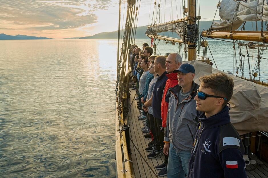 Grupa ludzi stoi ramię w ramię na pokładzie żaglowca, patrząc w stronę spokojnej tafli morza i zachodzącego słońca. W tle widać górzyste wybrzeże, co nadaje scenie podniosły i refleksyjny charakter.
