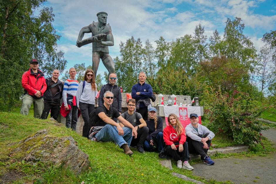Grupa osób pozuje do zdjęcia przed pomnikiem żołnierza z pociskiem artyleryjskim w rękach. Obok monumentu widoczny jest stół przykryty biało-czerwoną flagą z orłem oraz liczne znicze upamiętniające polskich bohaterów.
