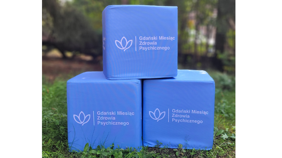 Trzy niebieskie kostki ustawione w plenerze, z logo w kształcie stylizowanego kwiatu oraz napisem „Gdański Miesiąc Zdrowia Psychicznego” na każdej z nich. Kostki stoją na trawie, a w tle widoczne są drzewa i rozmyte elementy ogrodu.