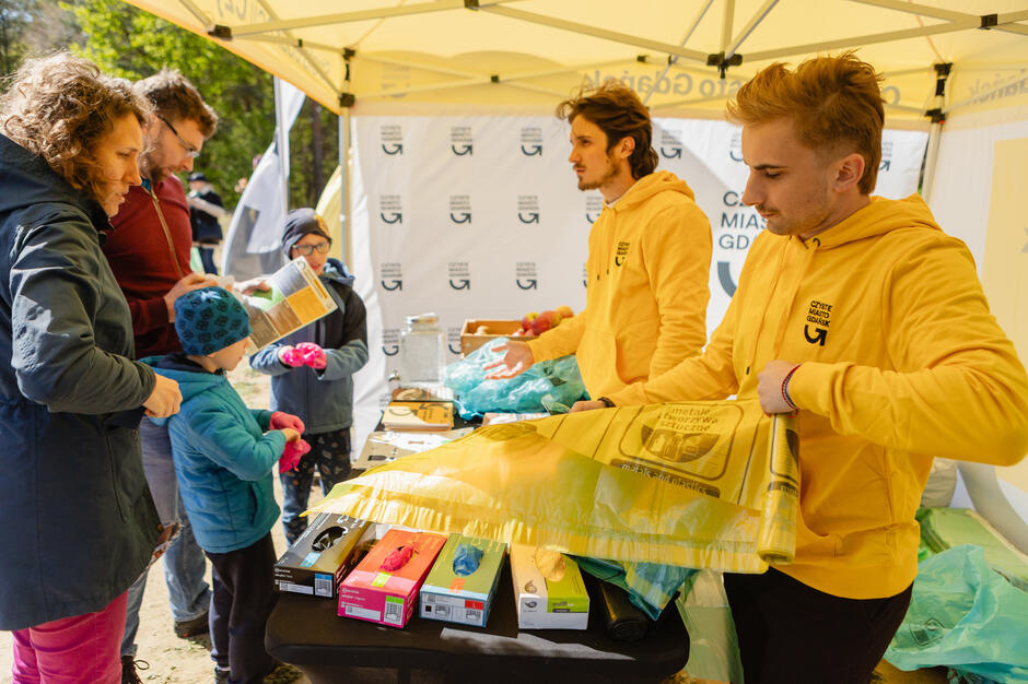 Pod namiotem akcji „Czyste Miasto Gdańsk” wolontariusze w żółtych bluzach rozdają worki i rękawiczki uczestnikom sprzątania. Przy stoisku stoi rodzina z dziećmi, przygotowując się do udziału w wydarzeniu ekologicznym.
