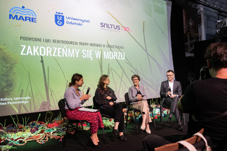 Trzy kobiety i mężczyzna siedzą na krzesłach podczas konferencji przed publicznością i rozmawiają.