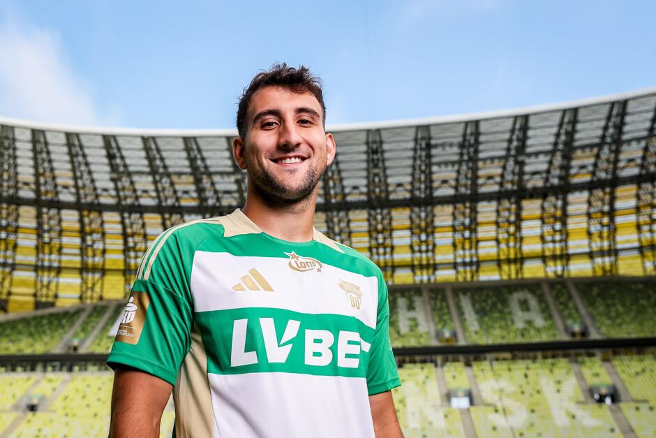 Młody mężczyzna w stroju piłkarskim pozuje do zdjęcia na tle trybun stadionu