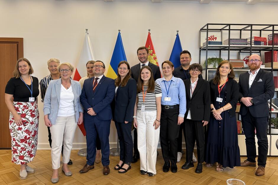 Wspólne pozowane zdjęcie 13 osób na tle stojących flag Polski, Ukrainy, Gdańska i Unii Europejskiej 