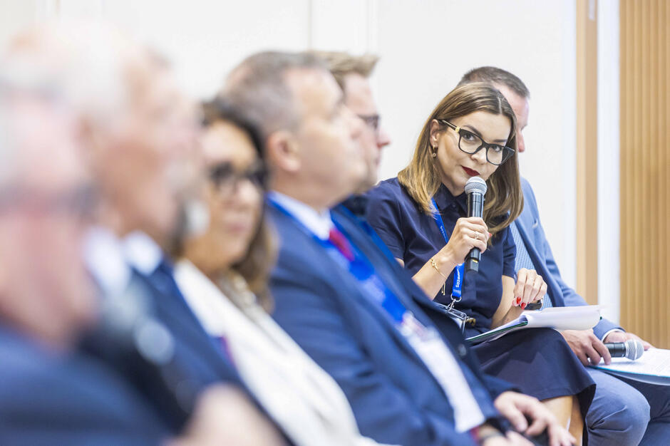 Na zdjęciu widać panel dyskusyjny, w którym uczestniczą osoby w formalnych strojach, a jedna z kobiet trzyma mikrofon i zabiera głos. Uczestnicy siedzą w rzędzie, koncentrując się na rozmowie w profesjonalnej atmosferze konferencyjnej. 