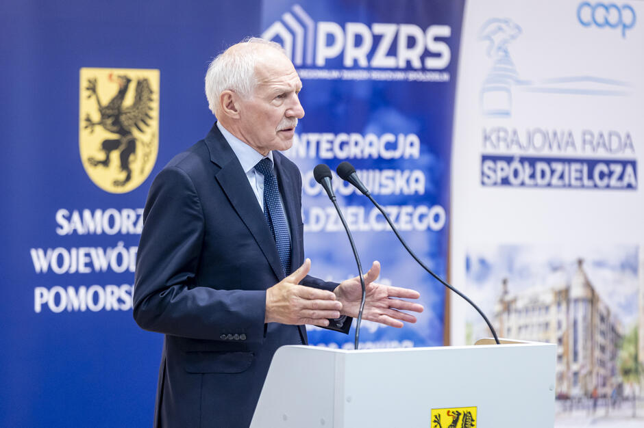 Na zdjęciu widać starszego mężczyznę w garniturze przemawiającego przy mównicy z dwoma mikrofonami. W tle znajdują się banery konferencyjne z logotypami instytucji, m.in. „Samorząd Województwa Pomorskiego” oraz „Krajowa Rada Spółdzielcza”. 