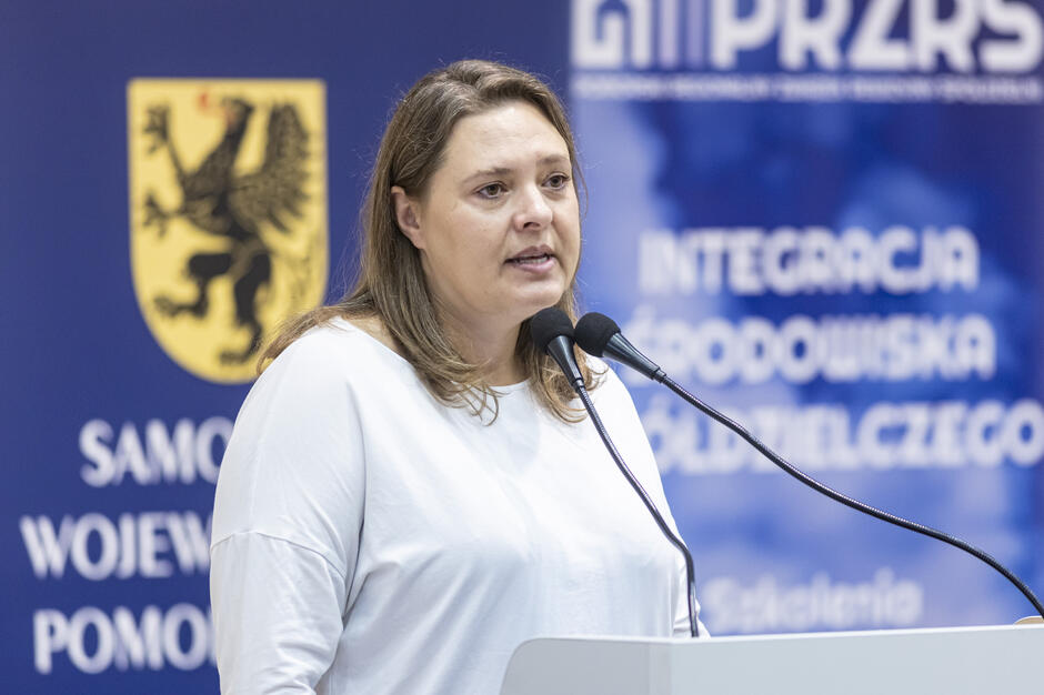 Na zdjęciu widać kobietę przemawiającą przy mównicy z dwoma mikrofonami, ubraną w białą bluzkę. W tle znajdują się banery konferencyjne z logotypami „Samorząd Województwa Pomorskiego” i „PRZRS”. 