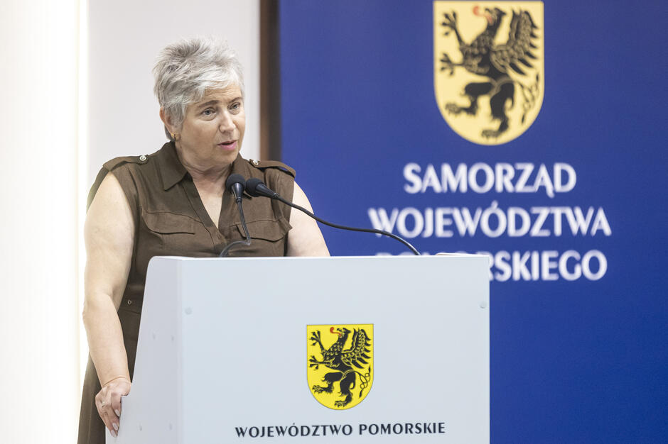 Na zdjęciu widać kobietę przemawiającą przy mównicy z dwoma mikrofonami, ubraną w białą bluzkę. W tle znajdują się banery konferencyjne z logotypami „Samorząd Województwa Pomorskiego” i „PRZRS”. 