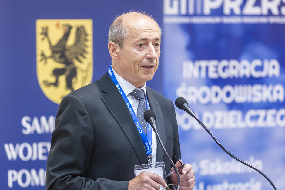 Na zdjęciu widać starszego mężczyznę w garniturze przemawiającego przy mównicy z dwoma mikrofonami. W tle znajdują się banery konferencyjne z logotypami instytucji, m.in. „Samorząd Województwa Pomorskiego” oraz „Krajowa Rada Spółdzielcza”. 