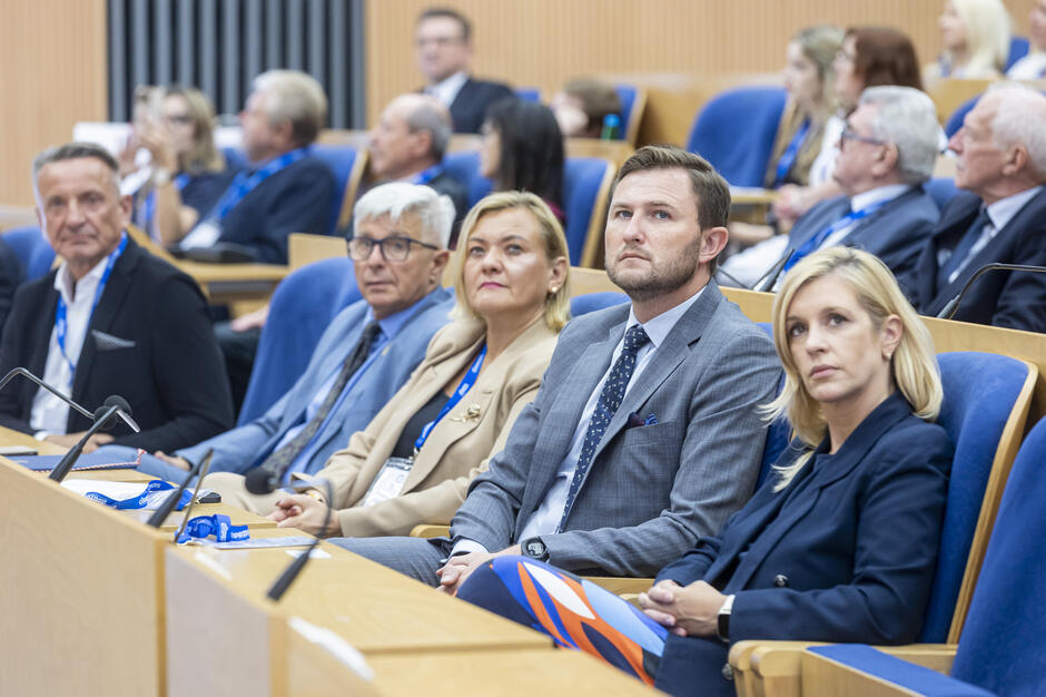 Na zdjęciu widać uczestników konferencji siedzących w sali audytoryjnej na niebieskich fotelach, uważnie słuchających wystąpień. W pierwszym rzędzie znajduje się pięć osób w formalnych strojach, patrzących w jedną stronę 