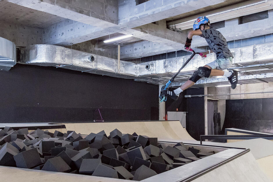 Chłopak w kasku skacze na hulajnodze i wpada do basenu z gąbkami w skateparku.