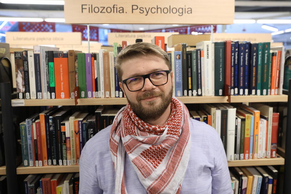 Na zdjęciu widać mężczyznę stojącego w bibliotece przed regałem z książkami z działu „Filozofia. Psychologia”. Ma okulary, brodę, jasne włosy i nosi jasną koszulę oraz czerwono-biały szalik.
