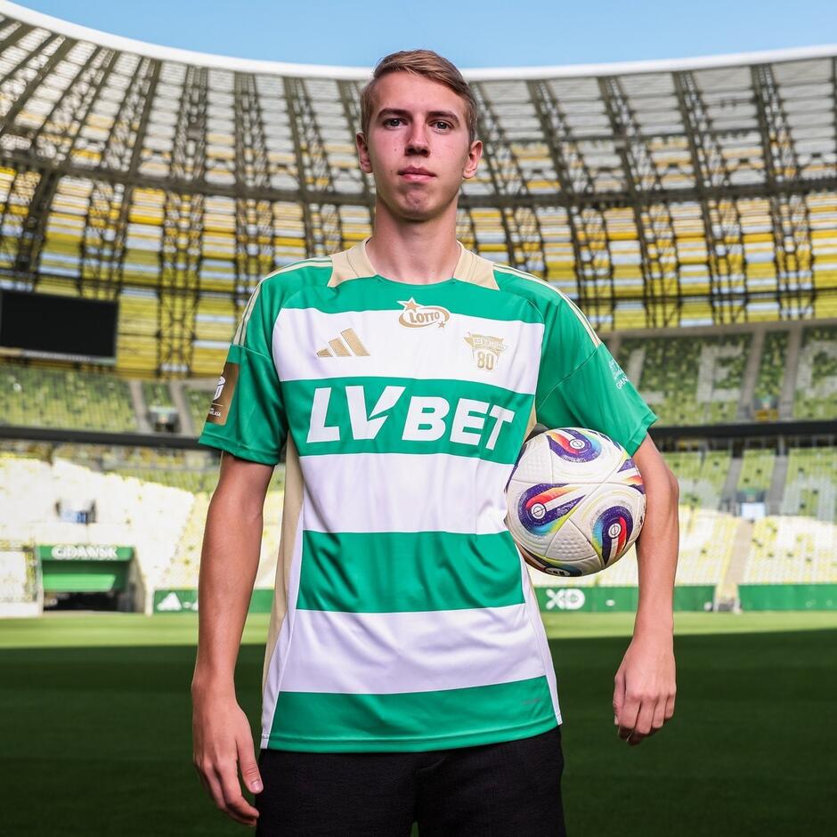 Zdjęcie młodego mężczyzny w biało-zielonym stroju piłkarskim, stoi na tle trybun stadionu Polsat Plus Arena Gdańsk