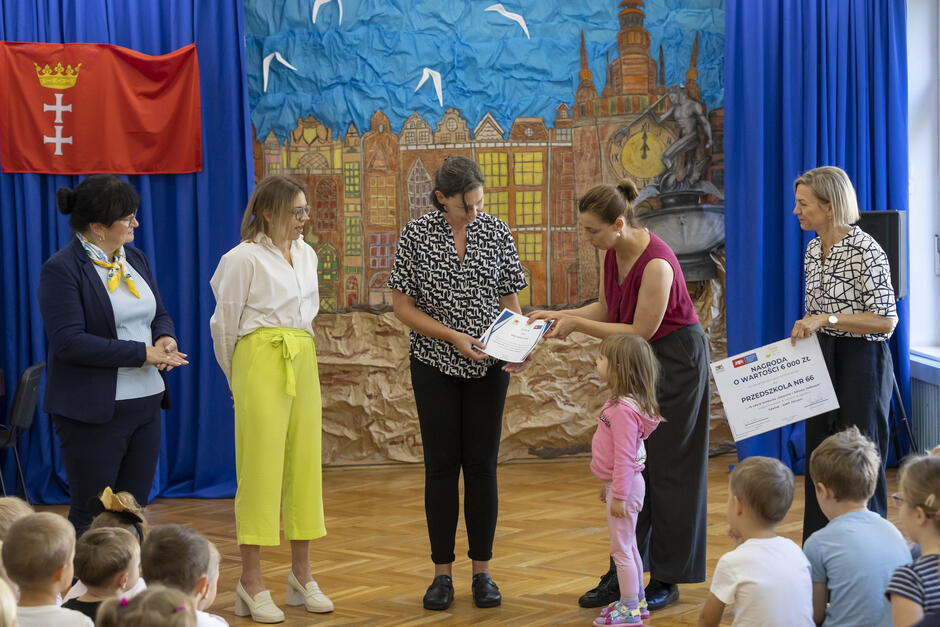 Pięć kobiet na scenie w sali przedszkola. Jedna kobieta wręcza drugiej dyplom. Przed nimi siedzą dzieci.