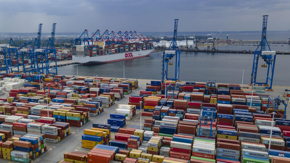 Na zdjęciu widzimy duży port kontenerowy. Na pierwszym planie znajdują się setki kolorowych kontenerów ustawionych w rzędach – należą one do różnych firm transportowych, takich jak Maersk, Hamburg Süd, CMA CGM czy Evergreen.