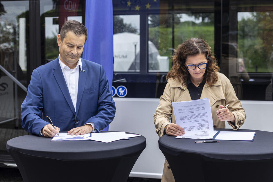 na zdjęciu kobieta i mężczyzna w średnim wieku, każde stoi przy wysokim stoliku i podpisuje umowę