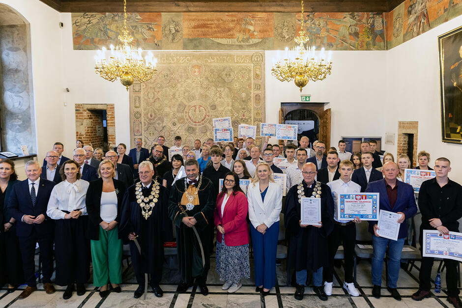 Duża grupa ludzi pozuje do fotografii w zdobnej sali.