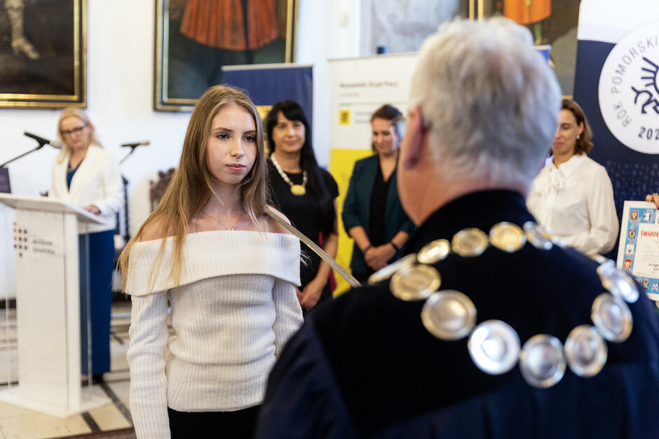 Młoda dziewczyna jest pasowana szablą na czeladniczkę w zdobnej sali.