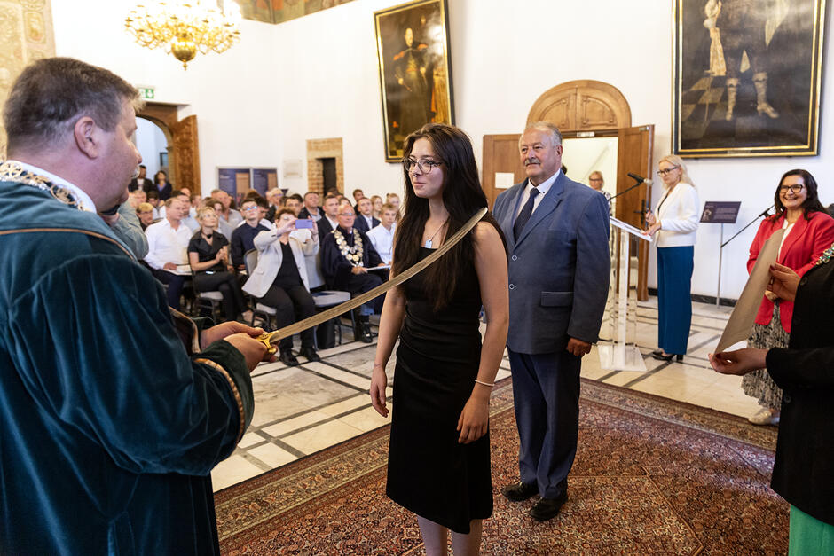 Mężczyzna w szatach pasuje szablą młoda kobietę na czeladnika w reprezentacyjnej sali.