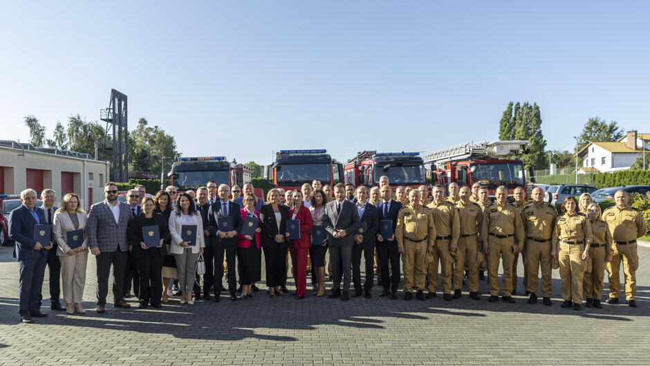 Grupa osób pozuje do zdjęcia ze strażakami na placu komendy straży pożarnej. Za nimi stoją wozy strażackie.