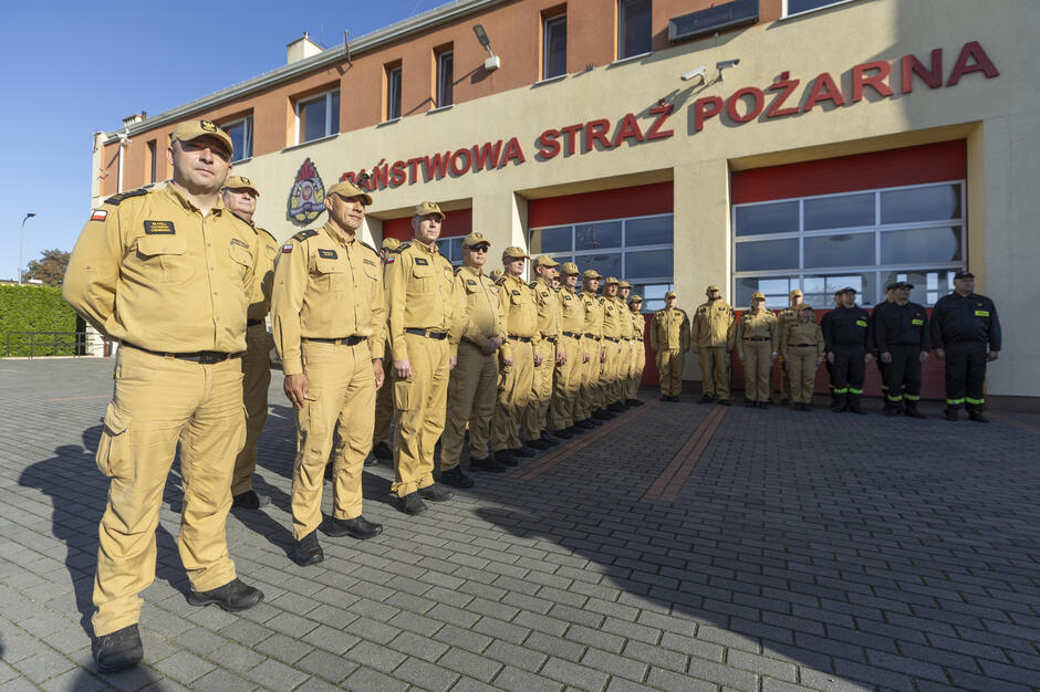 Strażacy stoją w szeregu przed remizą.
