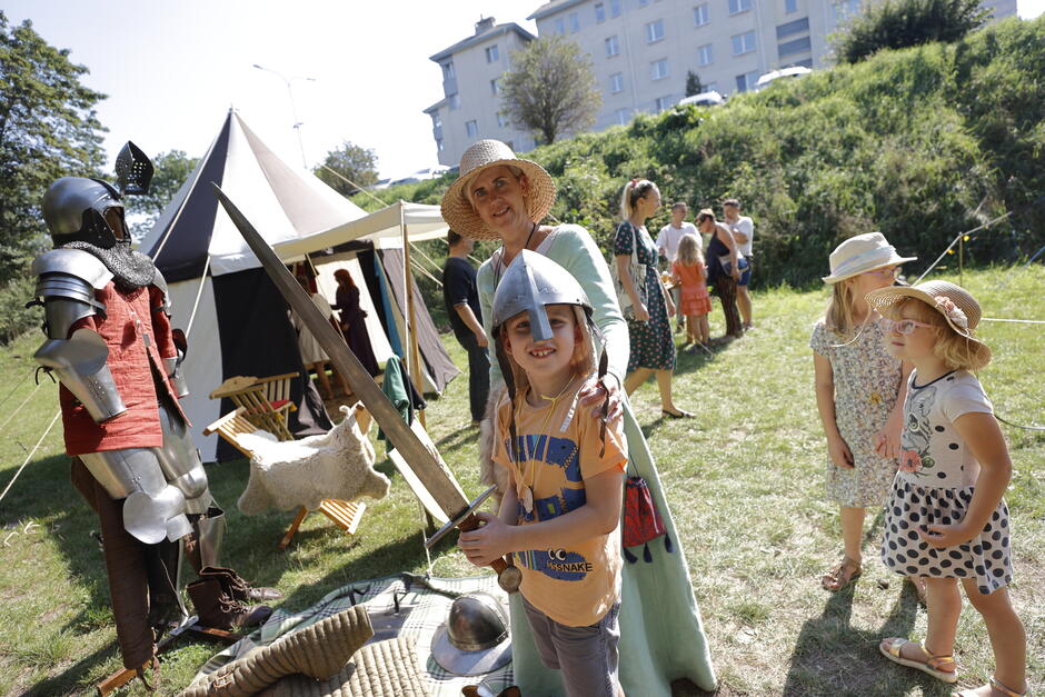 na zdjęciu chłopiec z rycerskim hełmem i mieczem, obok niego stoi babcia, w tle kilka innych osób stoi