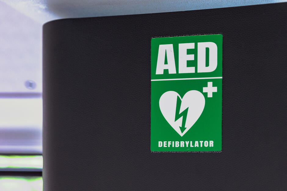 Na zdjęciu widać zieloną tabliczkę informacyjną z napisem **AED** i symbolem serca z błyskawicą, oznaczającą obecność defibrylatora. Taki znak wskazuje miejsce, w którym znajduje się automatyczny defibrylator zewnętrzny, używany w nagłych przypadkach zatrzymania krążenia. 