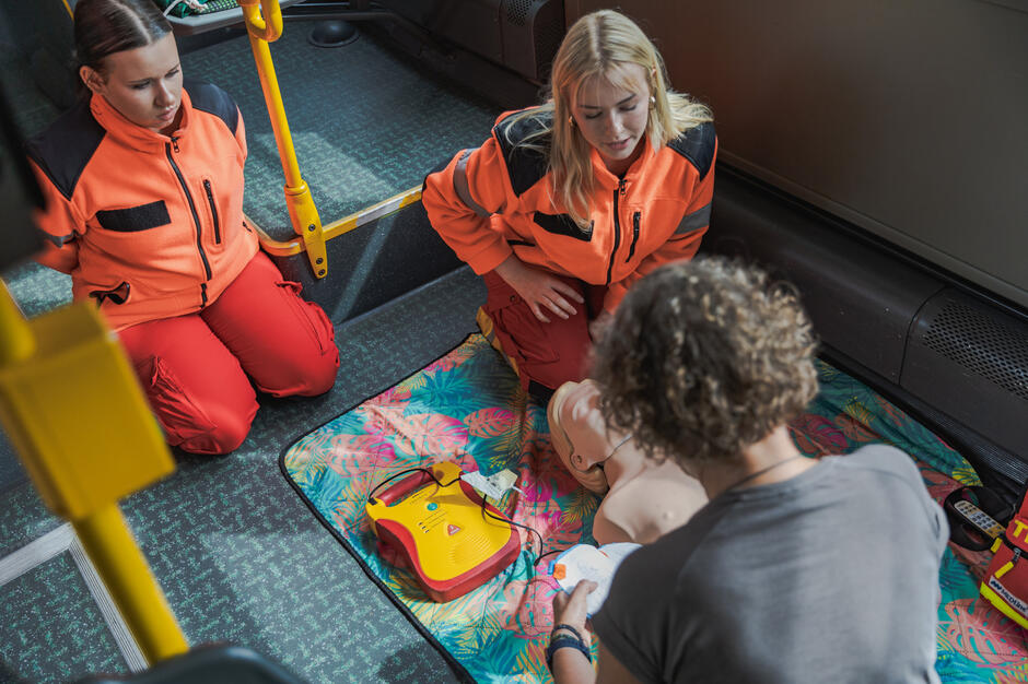 Na zdjęciu osoby w pomarańczowych strojach ratowniczych uczestniczą w szkoleniu z użycia defibrylatora AED na manekinie. Ćwiczenia odbywają się wewnątrz pojazdu, a sprzęt ratunkowy jest przygotowany obok manekina. 