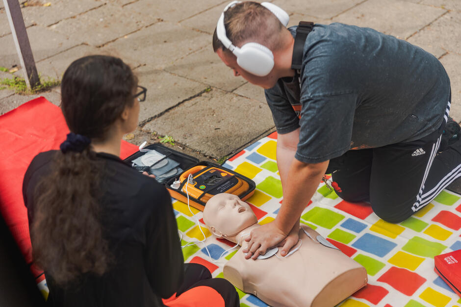 Na zdjęciu mężczyzna wykonuje uciski klatki piersiowej na manekinie, a obok znajduje się automatyczny defibrylator AED. Kobieta obserwuje przebieg ćwiczeń, co wskazuje na szkolenie z udzielania pierwszej pomocy. 