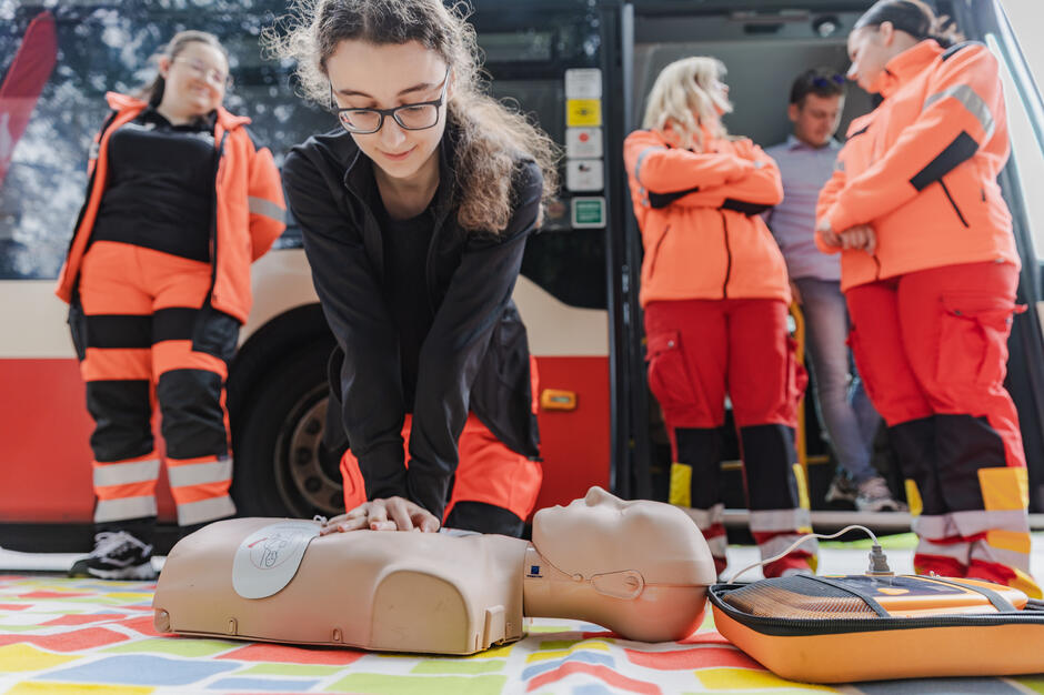 Na zdjęciu kobieta wykonuje uciski klatki piersiowej na manekinie podczas ćwiczeń z pierwszej pomocy. W tle stoją ratownicy medyczni obserwujący przebieg szkolenia. 