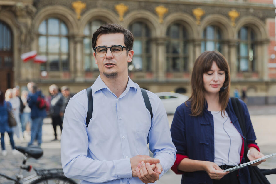Na zdjęciu znajduje się mężczyzna w okularach i koszuli oraz kobieta z mikrofonem nagłownym i mapą w ręku. Wygląda to na spacer miejski lub wydarzenie przewodnickie organizowane w przestrzeni publicznej.
