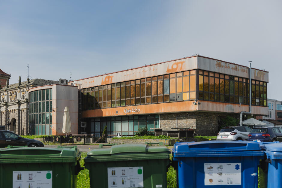 Na zdjęciu widoczny jest budynek z dużymi oknami i wyblakłym napisem „LOT”, w którym znajduje się „Club Echo”. Elewacja obiektu jest zniszczona i zardzewiała, a na pierwszym planie stoją pojemniki do segregacji odpadów.
