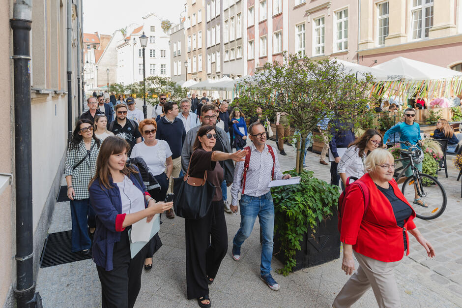 Na zdjęciu widać grupę osób spacerujących wąską uliczką w otoczeniu kamienic, prawdopodobnie podczas zorganizowanego wydarzenia lub wycieczki miejskiej. Część uczestników ma przy sobie notatki lub mapy, a jedna z kobiet wskazuje coś w kierunku dalszej części trasy.
