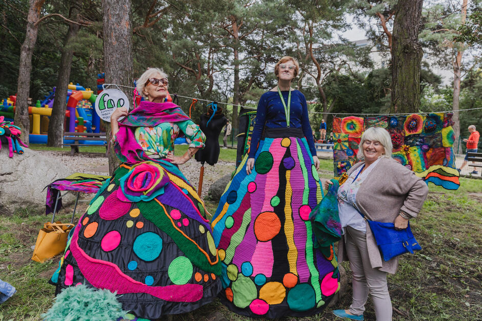 Dwie kobiety pozują do zdjęciach w wielkich, kolorowych spódnicach z włóczki i filcu w parku podczas festynu dzielnicowego.