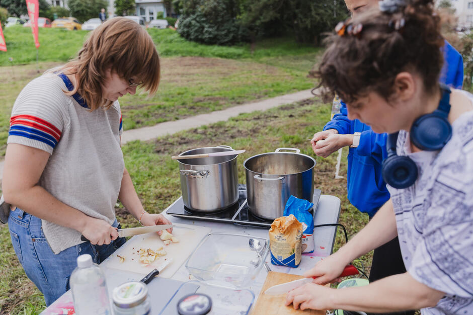 Dwie kobiety przy stole kroją jabłka, na stole stoją garnki, w których gotują się dżemy. Stół stoi w parku podczas festynu dzielnicowego.